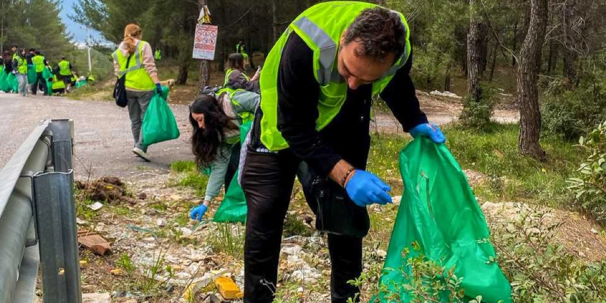 Ormanlık bir alanda, üzerlerinde neon sarı yelekler ve ellerinde mavi eldivenler bulunan gönüllüler, yeşil çöp poşetleri kullanarak çevre temizliği yapıyor. Ön planda eğilmiş bir şekilde çöp toplayan bir erkek ve hemen arkasında benzer şekilde çalışan bir kadın yer alıyor; arka planda ise yol kenarında temizliğe devam eden diğer gönüllüler görülüyor.