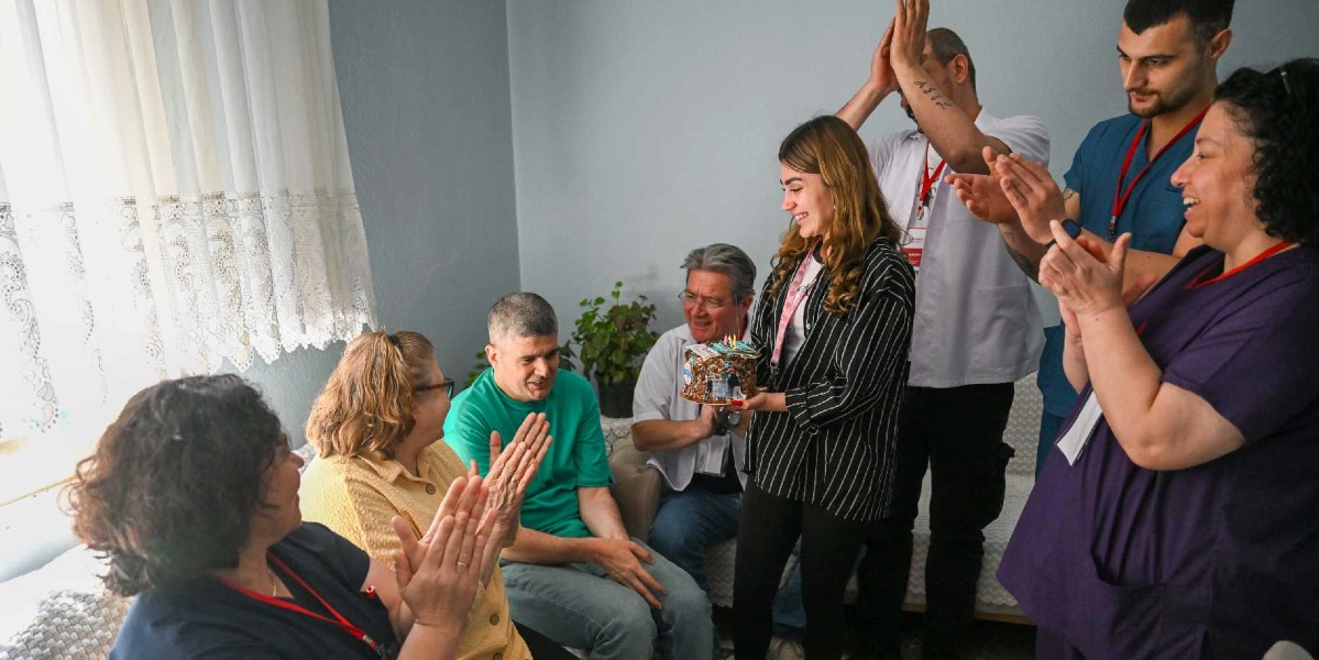 İç mekan bir odada, üzerinde mumlar yanan küçük bir pastayı tutan genç bir kadının etrafında toplanmış bir grup insan, neşeyle alkışlayarak doğum günü kutlaması yapıyor. Aralarında sağlık personeli veya bakım görevlisi oldukları anlaşılan, üniformalı kişilerin de bulunduğu grup, samimi bir ortamda bir araya gelmiş durumda.
