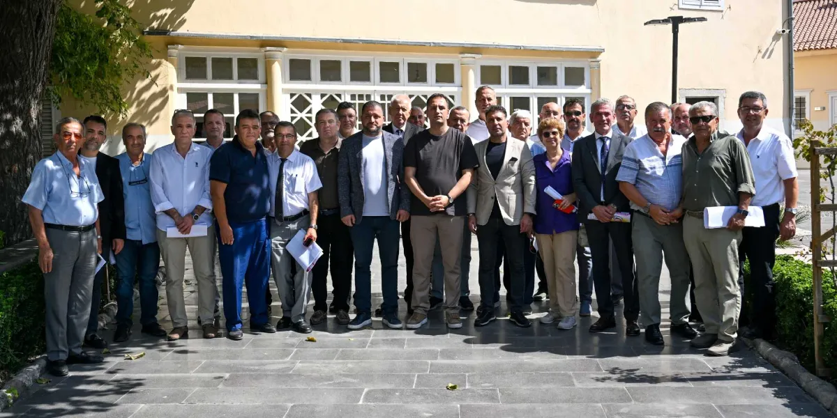 Dramalılar Köşkü’nün tarihi binası önünde yan yana dizilmiş, çoğunluğu erkeklerden oluşan yaklaşık yirmi kişilik bir grup, toplantı sonrası hatıra fotoğrafı çektiriyor. Katılımcıların bir kısmı takım elbiseli, bir kısmı ise günlük kıyafetler giymiş durumda; bazıları ellerinde belgeler tutuyor ve hepsi objektife bakarak gülümsüyor.