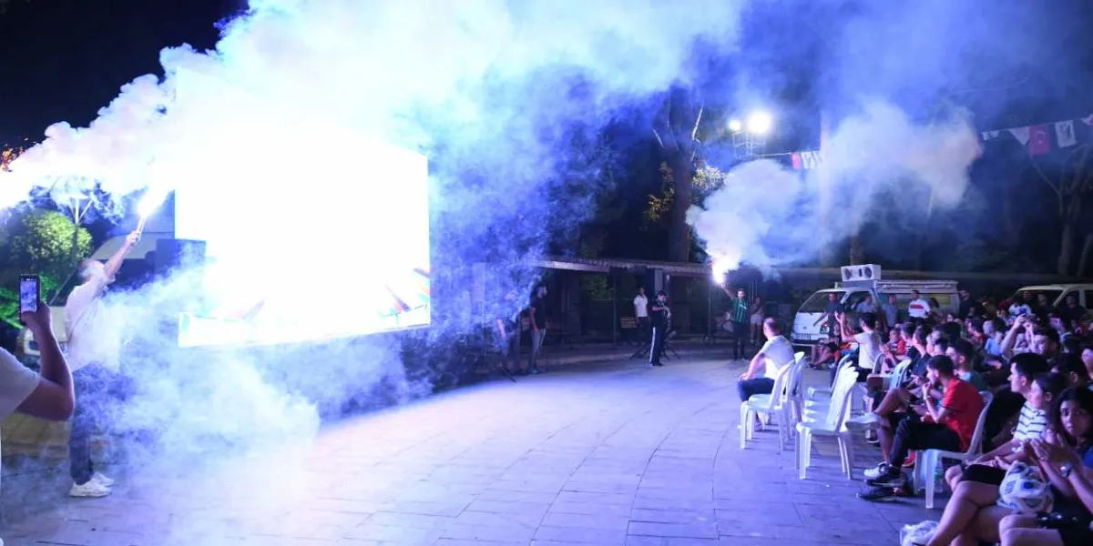 Gece vakti açık havada kurulan dev bir ekranın önünde toplanmış kalabalık, ellerinde yaktıkları renkli sis bombalarıyla kutlama yapıyor. Ekranın parlak ışığı ve yükselen yoğun dumanlar ortamı aydınlatırken, sandalyelerde oturan izleyiciler heyecanla maçı takip ediyor.