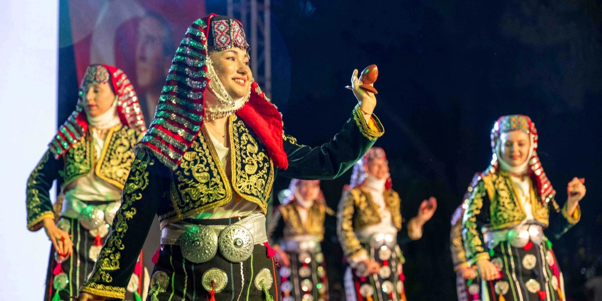 Geleneksel kıyafetler giymiş kadın dansçılar, ellerinde tuttukları ahşap kaşıklarla sahnede halk oyunu sergiliyor. Dansçılar, altın rengi işlemeli koyu yeşil ceketler, gümüş kemerler ve başlarında renkli süslemeleri olan örtülerle dikkat çekiyor. Ön plandaki dansçı, elindeki kaşığı havaya kaldırarak gülümseyen bir ifadeyle dansına devam ediyor.