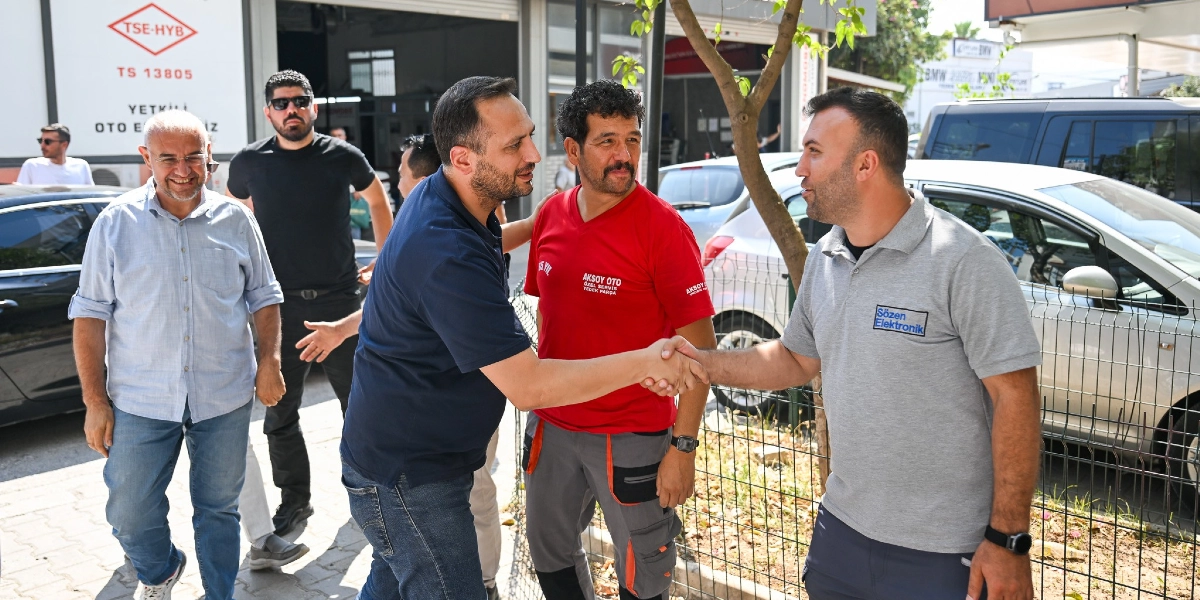 Bornova Belediye Başkanı Ömer Eşki, 3. Sanayi Sitesi'ndeki bir iş yerini ziyaret ederek çalışanlarla tokalaşıyor ve sohbet ediyor. Başkan Eşki lacivert polo yaka tişört ve kot pantolon giymiş, yanında ise kırmızı tişörtlü bir usta ve gri polo yaka tişörtlü bir çalışan yer alıyor. Arka planda sanayi sitesindeki dükkanlar, park halindeki araçlar ve TSE-HYB tabelası görülüyor.