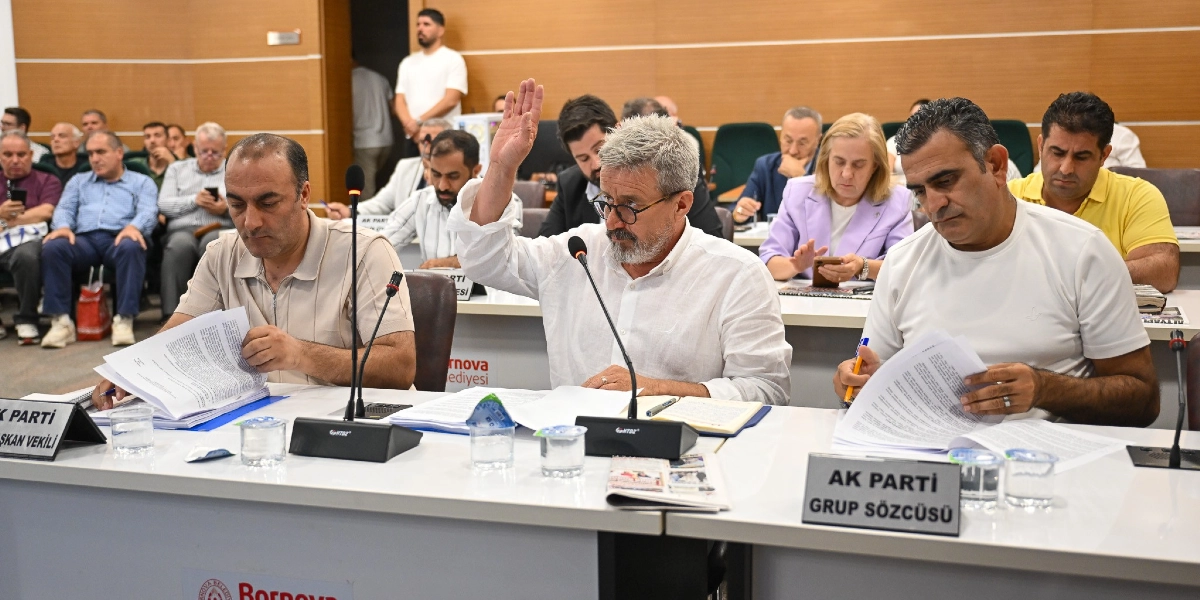Bornova Belediyesi meclis toplantısında, önlerinde AK Parti Grup Sözcüsü ve diğer görevlilere ait isimliklerin bulunduğu masada oturan üç erkek temsilci. Ortada oturan, beyaz gömlekli ve gözlüklü bir kişi elini kaldırarak söz istiyor; yanındaki diğer iki kişi ise önlerindeki belgeleri inceliyor. Arka planda toplantıya katılan diğer üyeler ve Bornova Belediyesi logosu görülüyor.