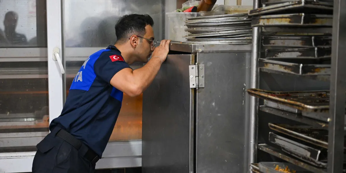 Üzerinde Türk bayrağı arması bulunan lacivert üniformalı bir zabıta memuru, bir fırın mutfağındaki metal dolabın içini dikkatle kontrol ediyor. Yan tarafta ise üst üste dizilmiş boş fırın tepsileri yer alıyor.