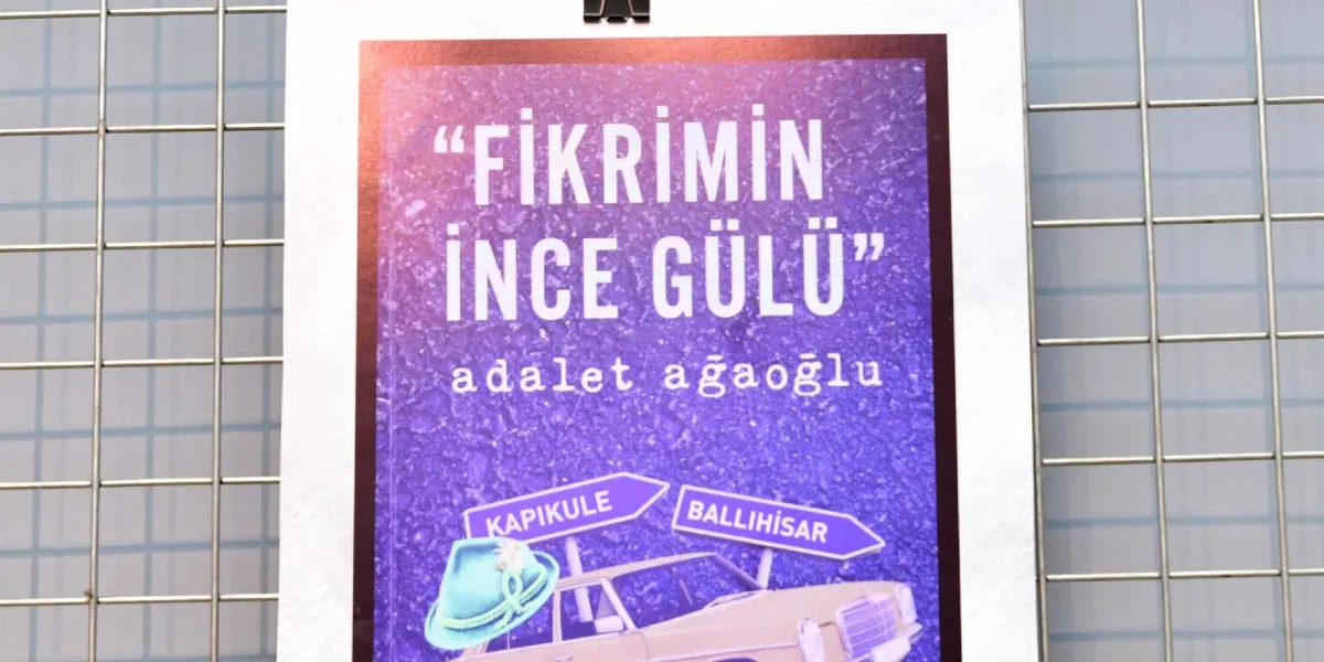 Metal bir ızgara üzerine asılmış mor renkli bir afişte, Adalet Ağaoğlu'nun Fikrimin İnce Gülü romanının kapağı yer alıyor. Kapak tasarımında, üzerinde Kapıkule ve Ballıhisar yazan iki yön tabelası, bir otomobil ve bir fötr şapka görseli bulunuyor.