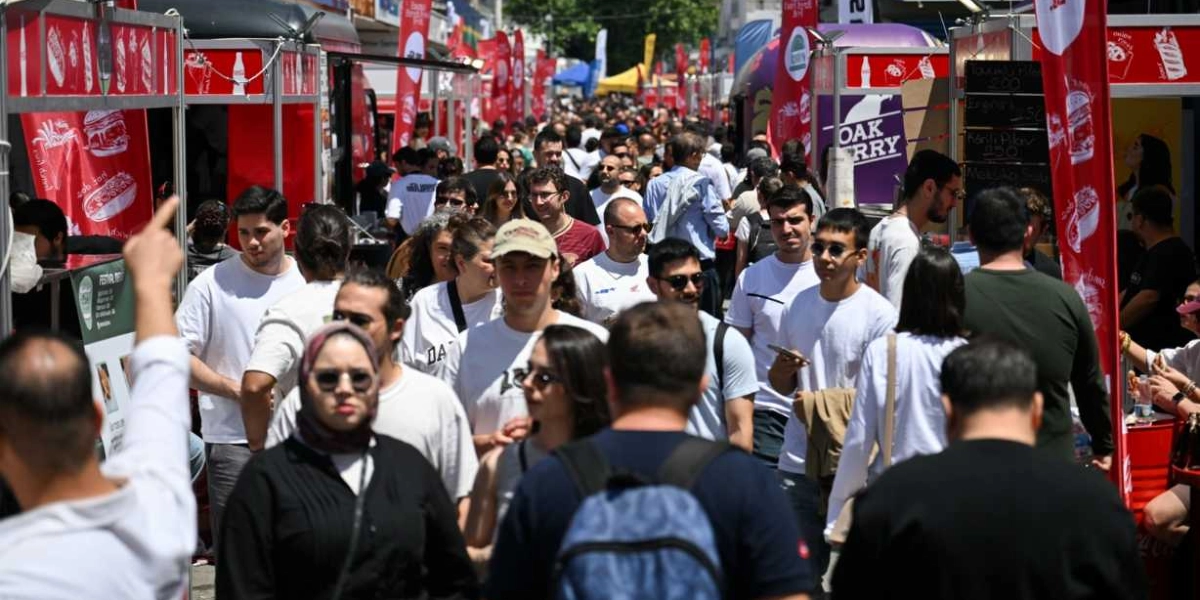 Bornova'da düzenlenen bir sokak festivalinde, yan yana dizilmiş yemek stantlarının önünde yürüyen ve alışveriş yapan kalabalık bir insan grubu. Gün ışığında çekilen fotoğrafta, stantların üzerinde kırmızı renkli reklam afişleri, menü panoları ve çeşitli yiyecek markalarının logoları yer alıyor. İnsanlar gündelik kıyafetleriyle stantlar arasında ilerlerken, ön planda bir kişinin eli bir yönü işaret ediyor.