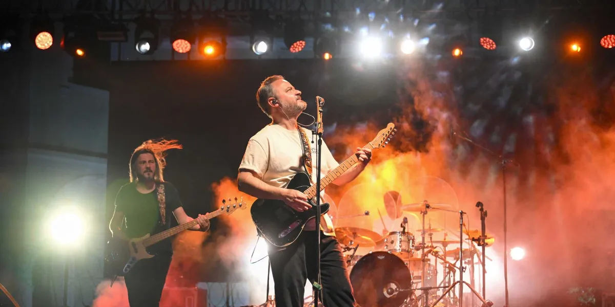 Sahne ışıkları ve yoğun duman altında konser veren bir müzik grubu. Ön planda krem rengi tişörtlü bir gitarist elinde elektro gitarıyla şarkı söylerken, sol tarafında uzun saçlı bir bas gitarist performans sergiliyor. Arka planda ise davul seti ve sahne aydınlatmaları yer alıyor.