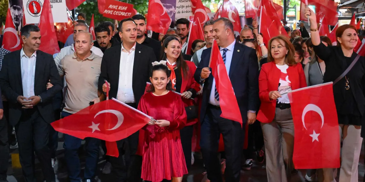 Bornova'da düzenlenen bir yürüyüşte, ellerinde Türk bayrakları taşıyan bir grup insan neşeyle ilerliyor. Grubun ön sırasında kırmızı kadife elbiseli genç bir kız, yanında takım elbiseli erkekler ve kırmızı ceketli kadınlar yer alıyor. Arka planda 