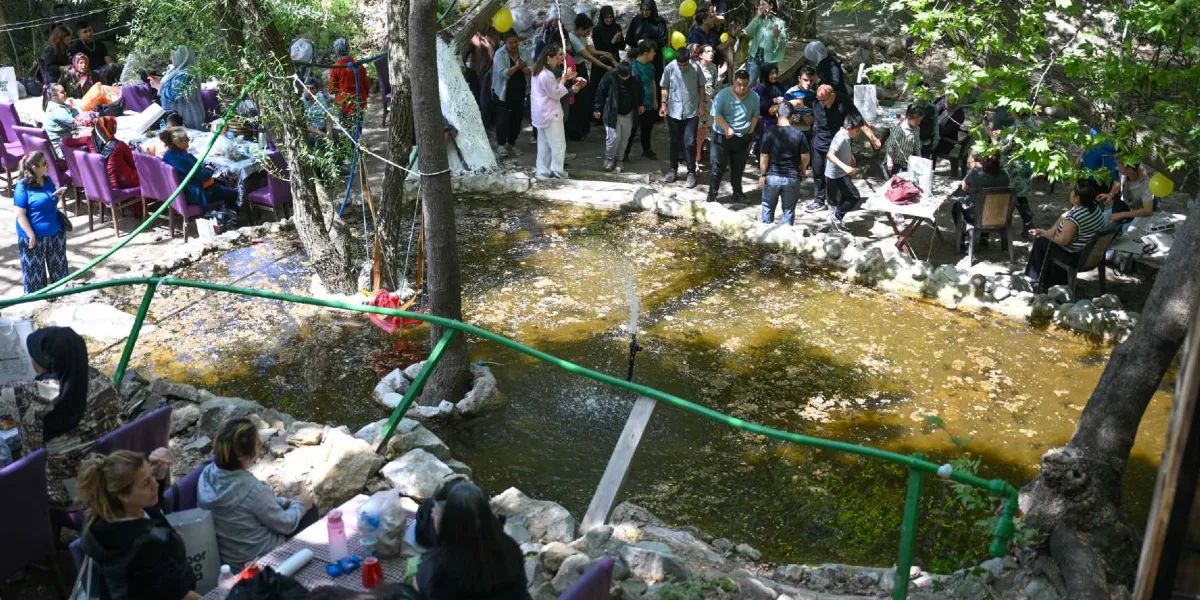 Ağaçlık ve gölgelik bir alanda, ortasında küçük bir süs havuzu bulunan bir bahçede toplanmış kalabalık bir grup insan. Havuzun etrafına yerleştirilmiş mor sandalyeler ve masalarda oturanlar, sohbet edenler ve ayakta duranlar görülüyor. Ortamda doğal bir atmosfer hakimken, insanlar bir arada vakit geçiriyor ve sosyalleşiyor.