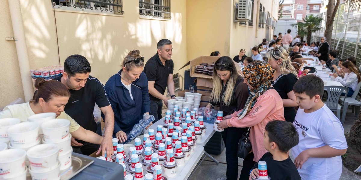 Açık havada kurulan uzun bir masanın etrafında toplanmış görevliler ve katılımcılar, plastik bardaklarda sunulan yiyecek ve içecekleri dağıtıyor. Ön planda, üzerinde çok sayıda paketli sütlü tatlı ve ayran bulunan masanın başında çalışanlar, gelen misafirlere ikramlarda bulunuyor. Arka planda ise masanın devamında oturan ve ayakta duran kalabalık bir grup insan, şenlik havasında bir arada vakit geçiriyor.