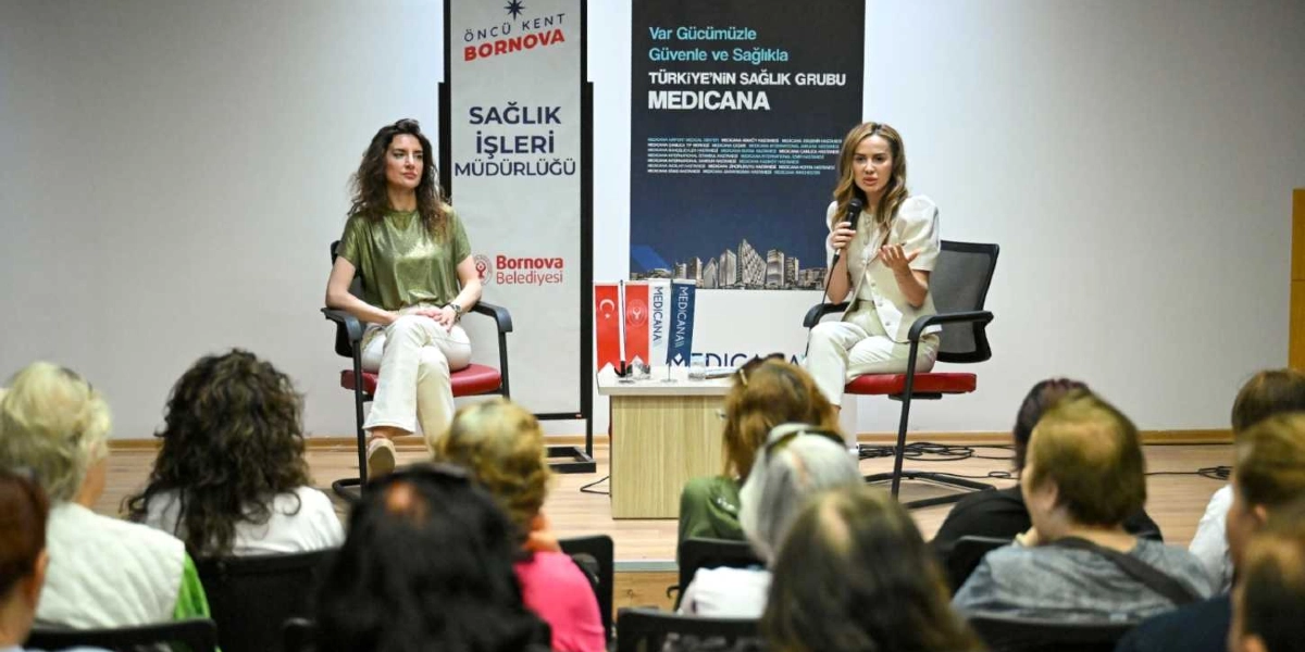 Bornova Belediyesi tarafından düzenlenen bir etkinlikte, iki kadın konuşmacı sahnedeki sandalyelerde oturarak katılımcılara hitap ediyor. Sağ taraftaki konuşmacı elinde mikrofonla sunum yaparken, sol taraftaki konuşmacı onu dinliyor. Arka planda Bornova Belediyesi Sağlık İşleri Müdürlüğü ve Medicana logolarının yer aldığı dikey afişler bulunuyor. İzleyiciler ise sahneye dönük bir şekilde oturarak konuşmayı takip ediyor.