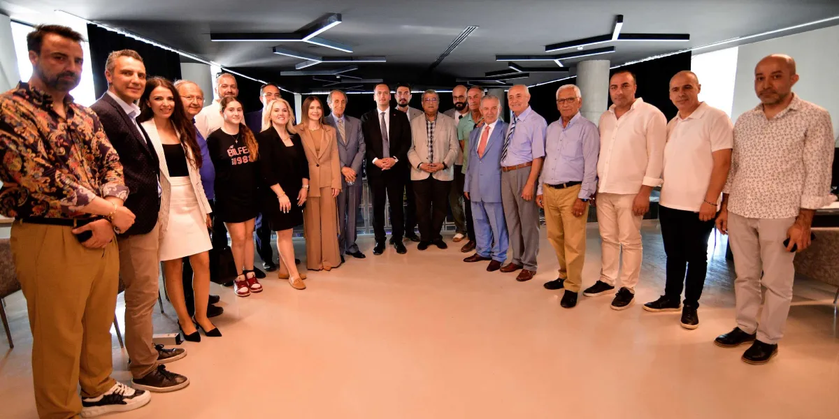Modern bir toplantı salonunda yan yana dizilmiş, farklı yaş ve giyim tarzlarına sahip yaklaşık yirmi kişilik bir grup, kameraya gülümseyerek poz veriyor. Grubun sol tarafında daha genç ve günlük kıyafetli kişiler, sağ tarafında ise genellikle takım elbise veya klasik pantolon-gömlek kombinleri tercih etmiş daha yaşlı bir grup yer alıyor. Arka planda tavanda geometrik siyah aydınlatmaların bulunduğu, geniş ve ferah bir iç mekan görülüyor.
