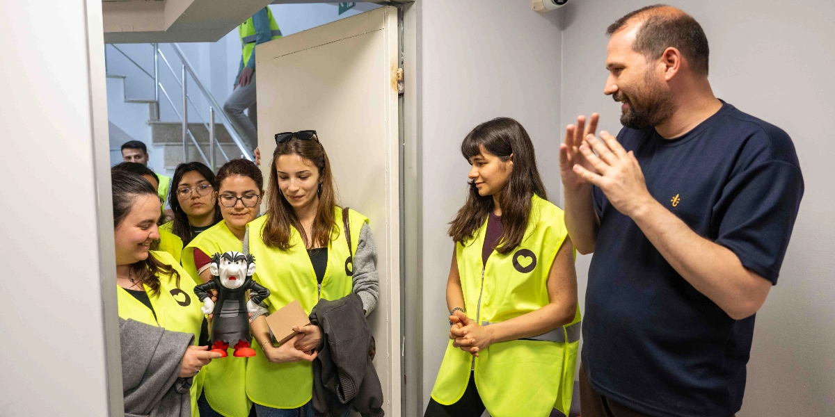 Üzerinde kalp logosu bulunan neon sarı yelekler giymiş bir grup genç, bir kapı girişinde sakallı bir adamla sohbet ediyor. Gençlerden biri elinde Gargamel karakterine benzeyen bir figür tutuyor. İç mekanda geçen bu buluşmada, katılımcılar gülümseyerek birbirlerini dinliyor.