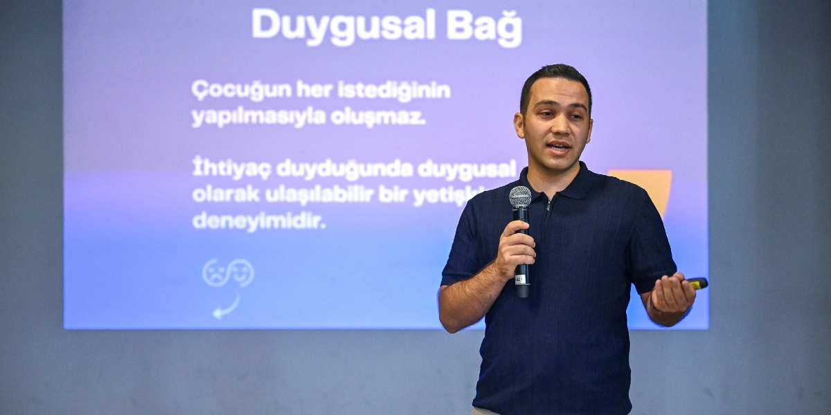 Koyu renkli polo yaka tişört giymiş bir konuşmacı, elinde mikrofonla bir sunum yaparken görülüyor. Arkasındaki yansıtılan ekranda 