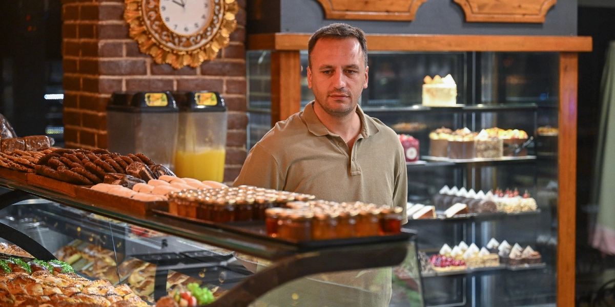 Bej renkli polo yaka tişört giymiş bir erkek, çeşitli hamur işleri ve tatlıların sergilendiği bir fırın tezgahının önünde duruyor. Arka planda tuğla duvar üzerinde bir saat ve raflarda dizili pastalar yer alıyor.