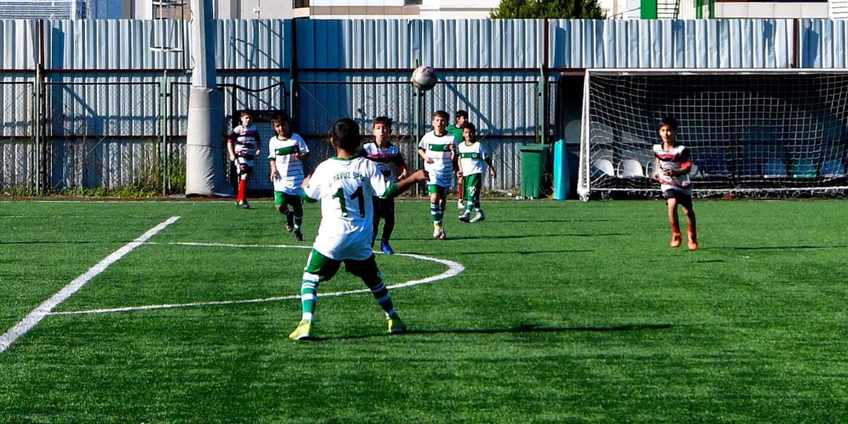 Yeşil çim sahada futbol oynayan çocuklardan oluşan bir grup. Ön planda, sırtında 11 numara ve Yavuz Spor yazılı beyaz formasıyla bir çocuk topa doğru yönelmiş durumda. Arka planda ise beyaz-yeşil formalar giymiş diğer oyuncular sahada koşuyor, havada bir futbol topu süzülüyor. Sahanın kenarında metal çitler ve bir kale direği bulunuyor.