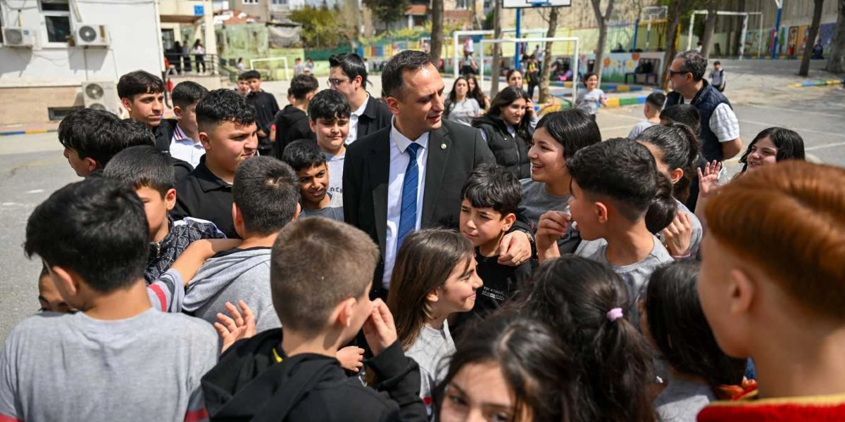 Takım elbiseli ve mavi kravatlı bir erkek, okul bahçesinde etrafını saran çok sayıda çocuk ve gençle bir arada duruyor. Çocukların bir kısmının omuzlarına elini koyarak onlarla samimi bir şekilde ilgilenen adam, öğrencilerin arasında gülümsüyor. Arka planda basketbol potası ve okul binası bulunan açık hava ortamında, öğrenciler meraklı ve mutlu bir ifadeyle adamın etrafında toplanmış durumda.