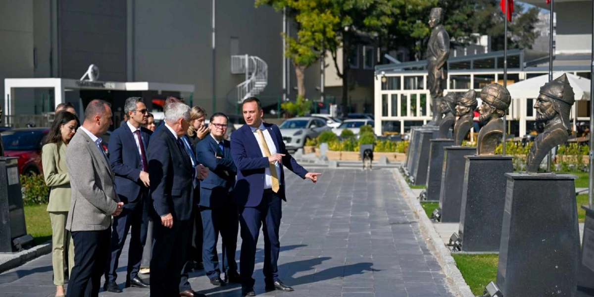 Açık havada, taş döşeli bir yolda yürüyen bir grup resmi giyimli kişi, sağ tarafta sıralanmış olan tarihi figürlerin büstlerini incelemektedir. Grubun önünde yer alan ve lacivert takım elbiseli, sarı kravatlı bir kişi, eliyle büstleri işaret ederek yanındakilere bilgi vermektedir. Arka planda ise ağaçlar, park halindeki araçlar ve modern bir bina yapısı görülmektedir.