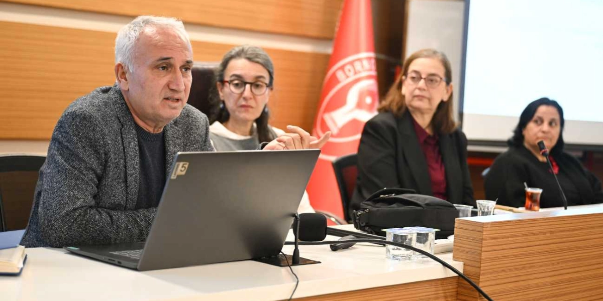 Toplantı masasında oturan dört kişilik bir grup, Bornova Belediyesi'nin logosunun bulunduğu kırmızı bir bayrağın önünde sunum yapıyor. Ön planda gri ceketli, beyaz saçlı bir erkek konuşmacı dizüstü bilgisayarının başında yer alıyor. Onun yanında, arkasında ve sağ tarafında ise üç kadın katılımcı dikkatle dinliyor. Masanın üzerinde mikrofonlar, su bardakları ve çay bardağı bulunuyor.