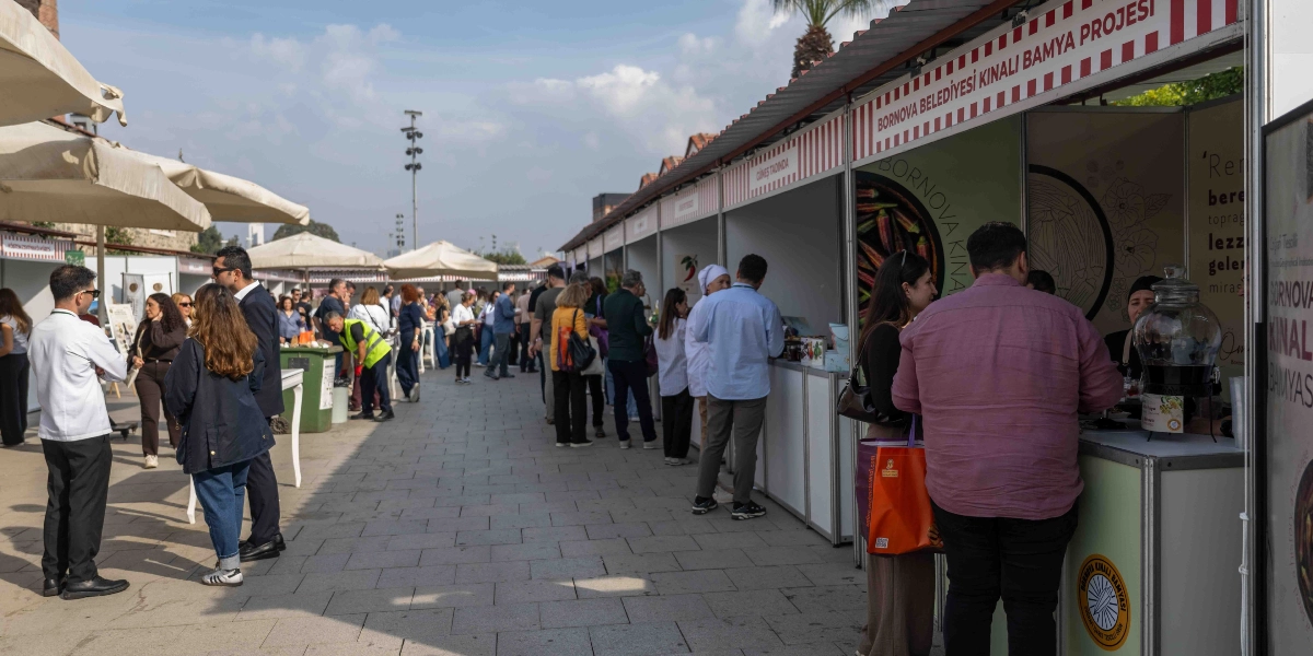 Açık havada kurulan bir festival alanında, yan yana dizilmiş beyaz stantların önünde insanlar geziniyor. Sağ tarafta yer alan 