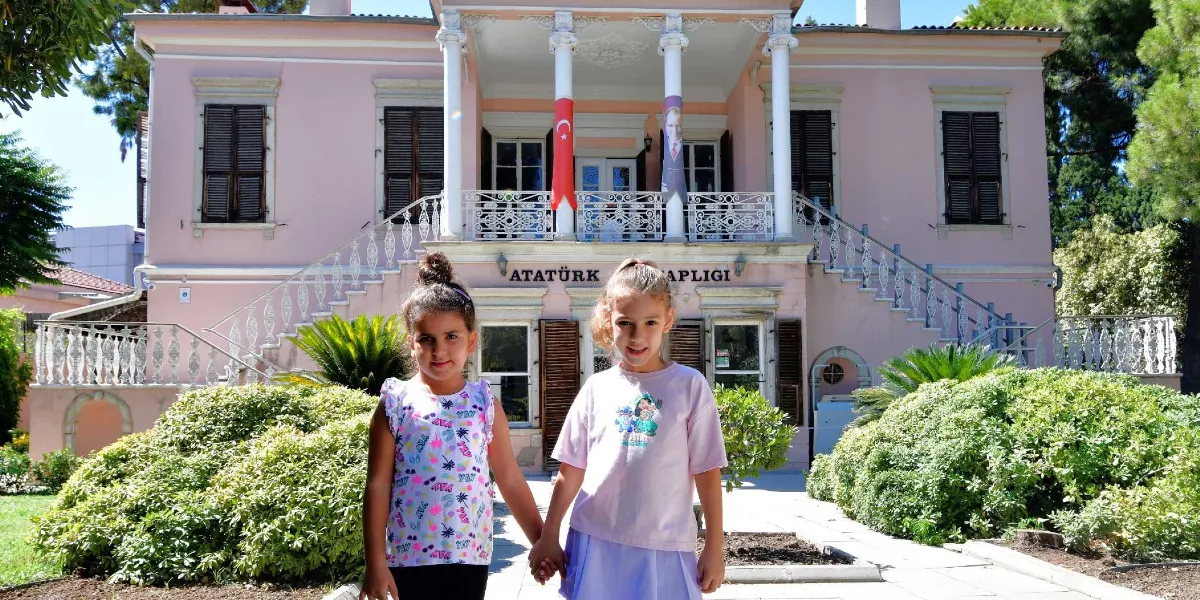 Bornova'daki tarihi pembe renkli Atatürk Kitaplığı binasının önünde el ele tutuşan iki kız çocuğu gülümsüyor. Arka planda sütunlu girişi, Türk bayrağı ve Atatürk posteriyle dikkat çeken, bahçeli ve iki katlı tarihi bina yer alıyor.
