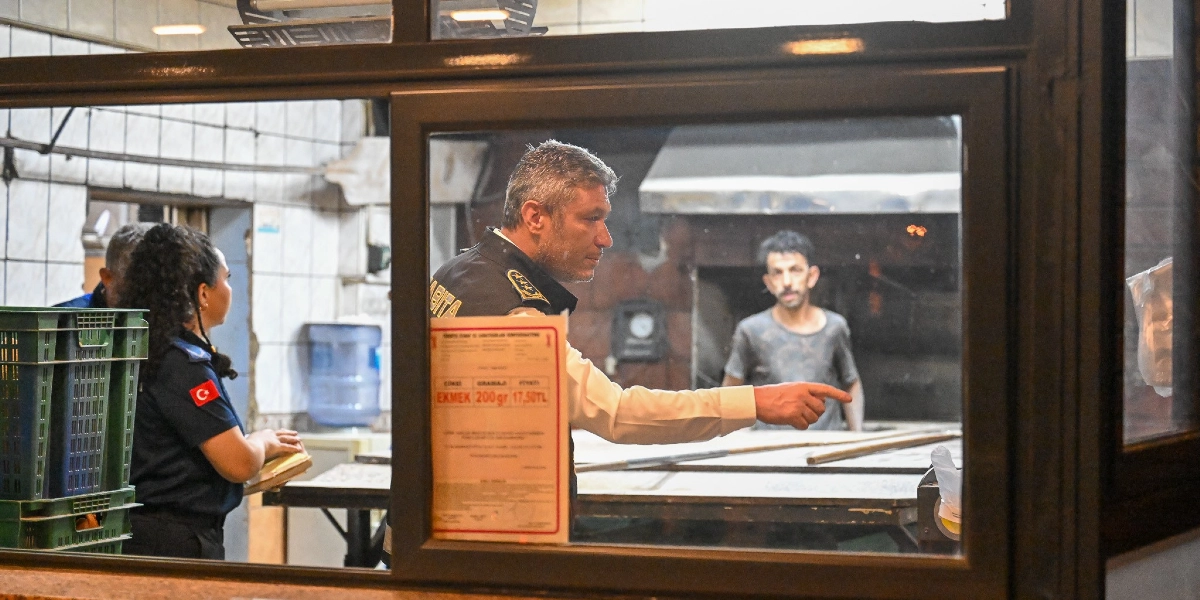 Zabıta üniformalı bir yetkili, bir fırının üretim alanında eliyle bir noktayı işaret ederek denetim yapıyor. Yanında zabıta üniformalı bir kadın personel not tutarken, arka planda fırın çalışanı tezgah başında duruyor. Ön plandaki camın üzerinde, ekmek gramajı ve fiyat bilgisinin yer aldığı resmi bir tarife belgesi asılı duruyor.