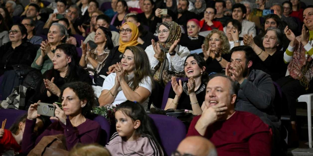 Kapalı bir salonda, mor koltuklarda oturan ve büyük bir ilgiyle izledikleri gösteriyi alkışlayan, aralarında çocukların da bulunduğu kalabalık bir izleyici grubu. Ön planda, elinde telefonuyla çekim yapan bir kadın, yanında oturan küçük bir kız çocuğu ve gülümseyerek alkışlayan izleyiciler yer alıyor.