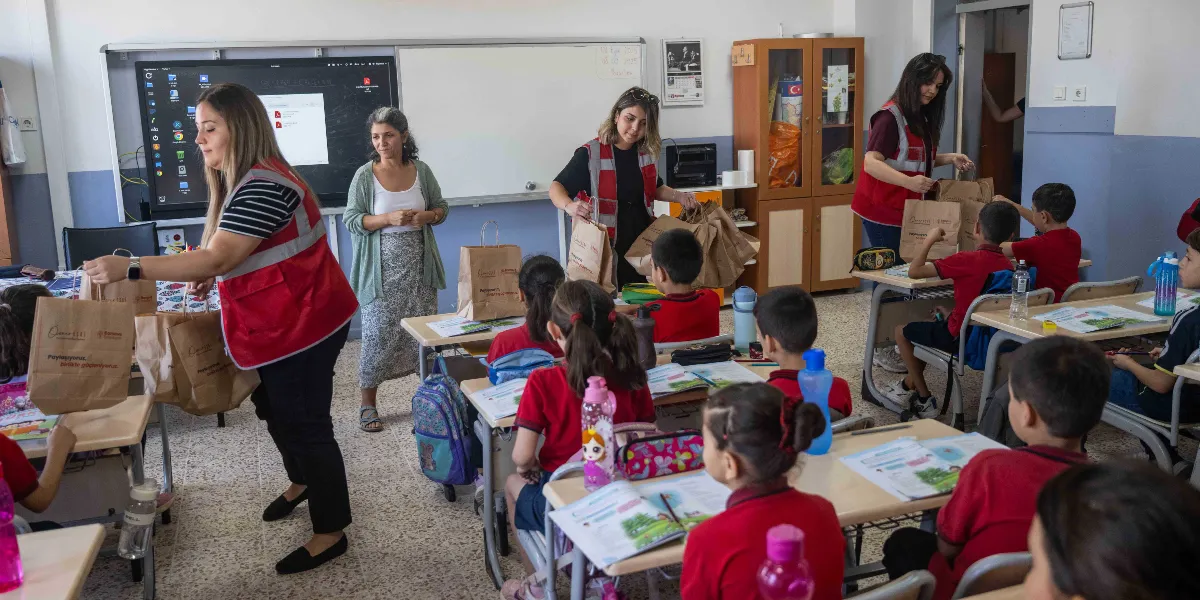 Sınıf ortamında, üzerinde Bornova Belediyesi logolu kırmızı yelekler bulunan görevliler, sıralarında oturan ilkokul öğrencilerine içinde kırtasiye malzemeleri bulunan hediye çantalarını dağıtıyor. Öğrenciler kırmızı okul formalarıyla sıralarında otururken, görevliler sınıfta dolaşarak çantaları öğrencilere veriyor. Arka planda akıllı tahta, dolaplar ve ayakta duran bir öğretmen bulunuyor.