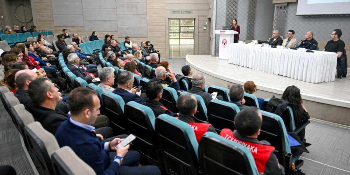 Modern bir konferans salonunda düzenlenen deprem konulu panelde, kürsüde konuşma yapan bir kadın ve masada oturan dört panelist yer alıyor. Salonun izleyici kısmında, Bornova Belediyesi logolu kürsünün önünde ve yanındaki masada uzmanlar bulunurken, dinleyiciler arasında üzerinde 