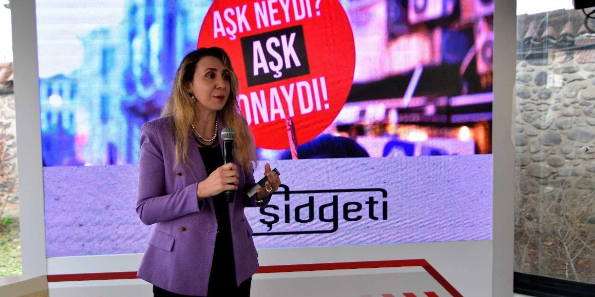Lila renkli ceket giymiş bir kadın, elinde mikrofonla konuşma yapıyor. Arkasındaki büyük ekranda kırmızı bir daire içinde 