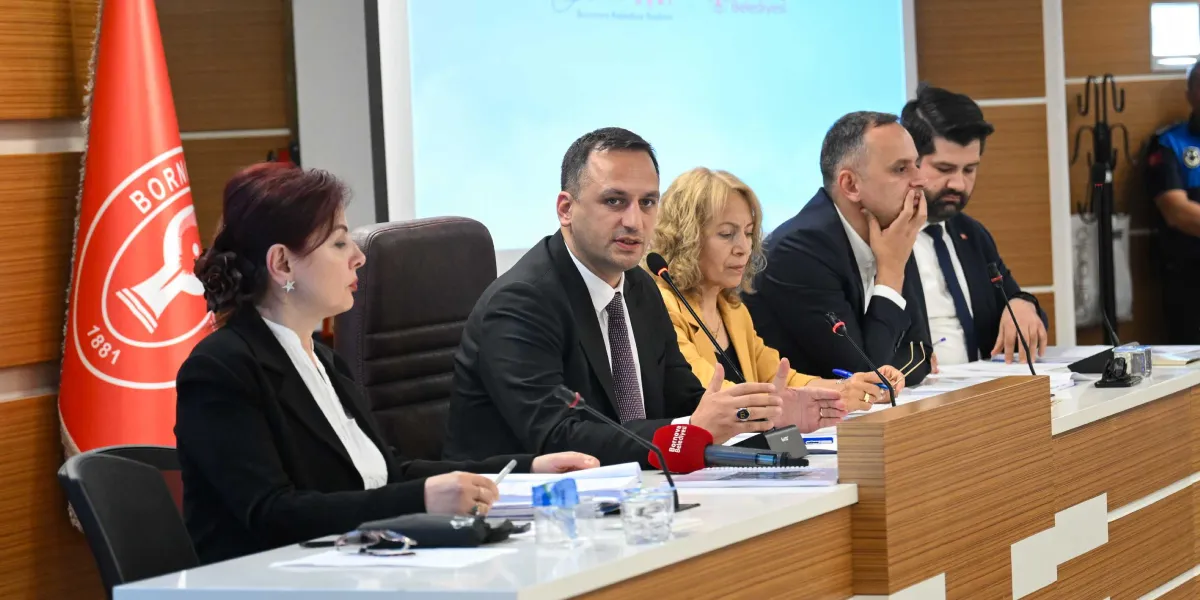 Bornova Belediyesi meclis salonunda, üzerinde Bornova Belediyesi logosu bulunan kırmızı bir bayrağın yanında, bir masada oturan dört kişilik bir heyet toplantı yapıyor. Masanın merkezinde konuşma yapan takım elbiseli bir erkek, yanında sarı ceketli bir kadın ve diğer iki takım elbiseli erkek yer alıyor. Önlerinde mikrofonlar, belgeler ve su bardakları bulunan heyet, resmi bir oturum gerçekleştiriyor.