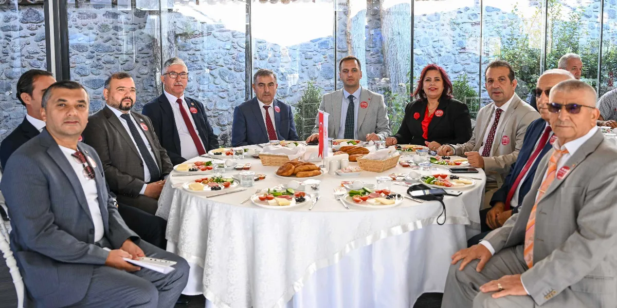 Beyaz örtülü yuvarlak bir masa etrafında toplanmış, çoğunluğu takım elbiseli erkeklerden ve bir kadından oluşan grup, kahvaltı eşliğinde bir toplantı gerçekleştiriyor. Katılımcıların yakalarında kırmızı renkli, üzerinde yazı bulunan rozetler dikkat çekiyor. Arka planda taş duvarlı ve cam bölmeli aydınlık bir mekan yer alıyor.