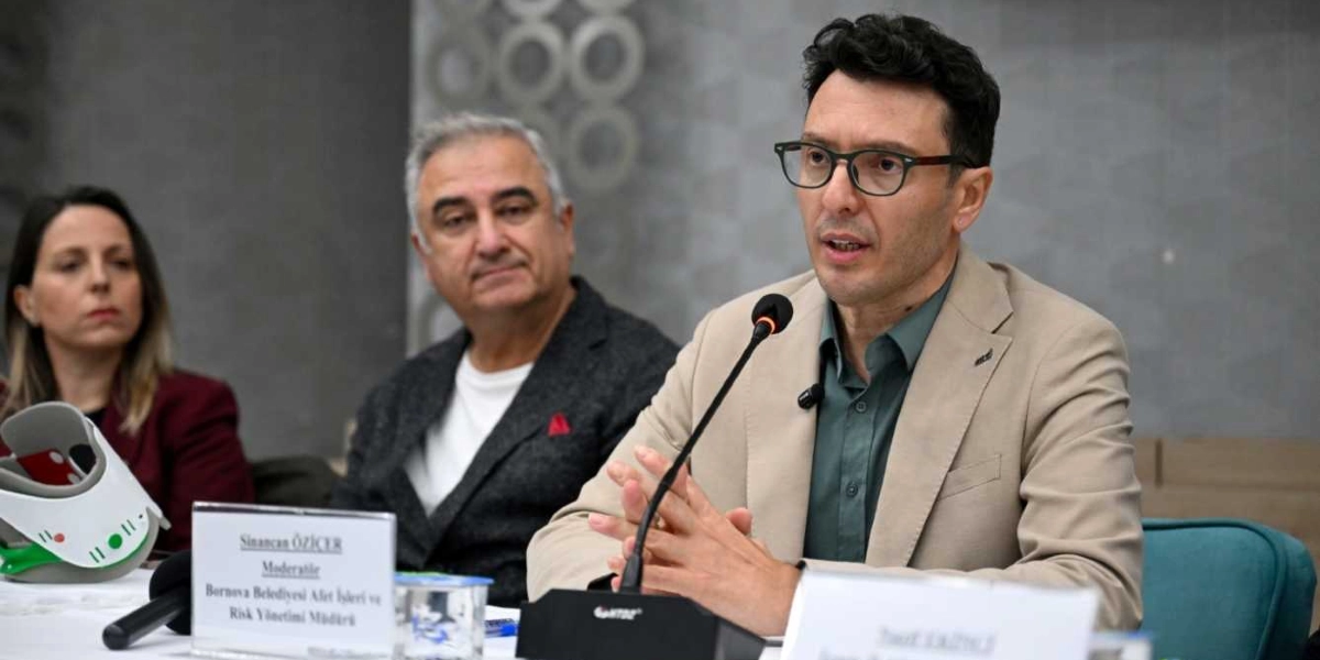 Bir panelde konuşma yapan gözlüklü bir erkek, yanında oturan diğer katılımcılarla birlikte masada yer alıyor. Masanın üzerinde 