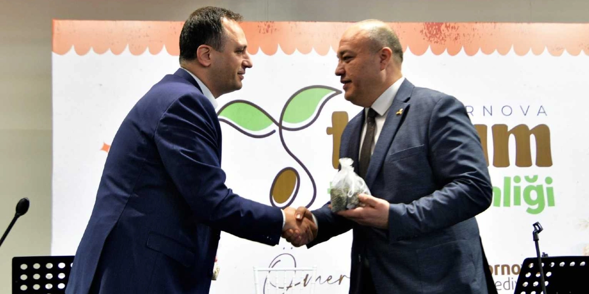 Lacivert takım elbiseli iki erkek, arka planında Bornova Tohum Takas Şenliği yazılı bir afişin önünde tokalaşıyor. Sağdaki kişi, elinde küçük, şeffaf bir tohum kesesi tutuyor.