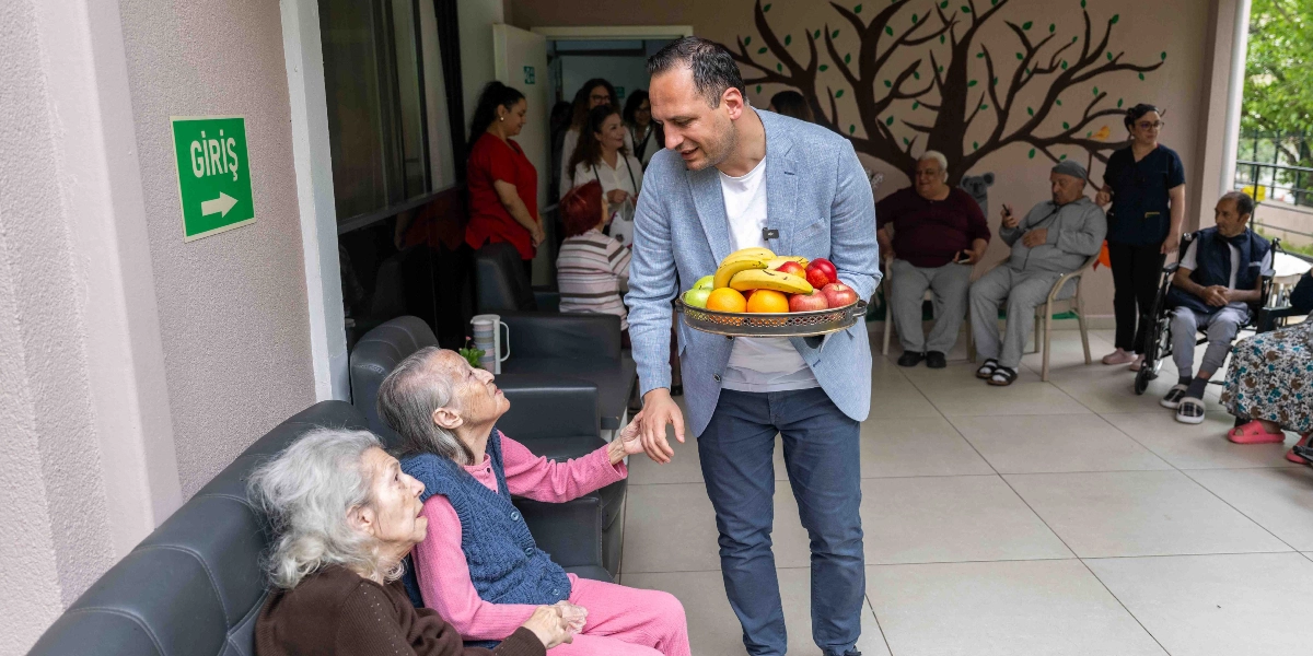 Açık mavi ceketli bir erkek, bir huzurevi veya bakım merkezinde elinde meyve dolu bir tepsiyle yaşlı kadınların elini tutarak onlarla sohbet ediyor. Arka planda duvarında ağaç figürü bulunan bir salonda oturan diğer yaşlılar ve görevliler yer alıyor. Duvarın üzerinde yeşil renkli, beyaz yazılı bir 