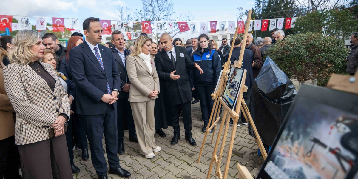 Açık havada düzenlenen bir anma töreninde, aralarında Bornova Belediye Başkanı Mustafa İduğ'un da bulunduğu bir grup yetkili, ahşap şövale üzerine yerleştirilmiş Türkan Bebek'i temsil eden bir resmi incelemektedir. Katılımcılar resmi ve çevresindeki alanı dikkatle izlerken, arka planda Türk bayrakları ve Atatürk posterleriyle süslenmiş bir alan görülmektedir.