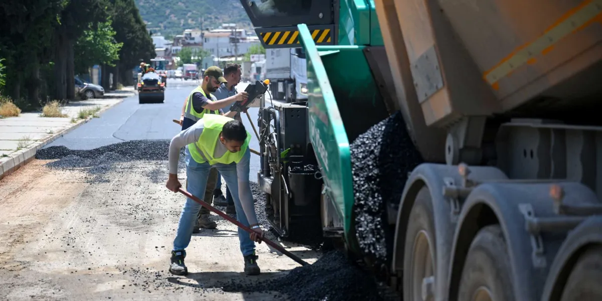 Pınar Caddesi'nde yol yenileme çalışmaları kapsamında, üzerinde reflektörlü yelek bulunan işçiler asfalt serme makinesi ve kamyon yardımıyla yola sıcak asfalt döküyor.