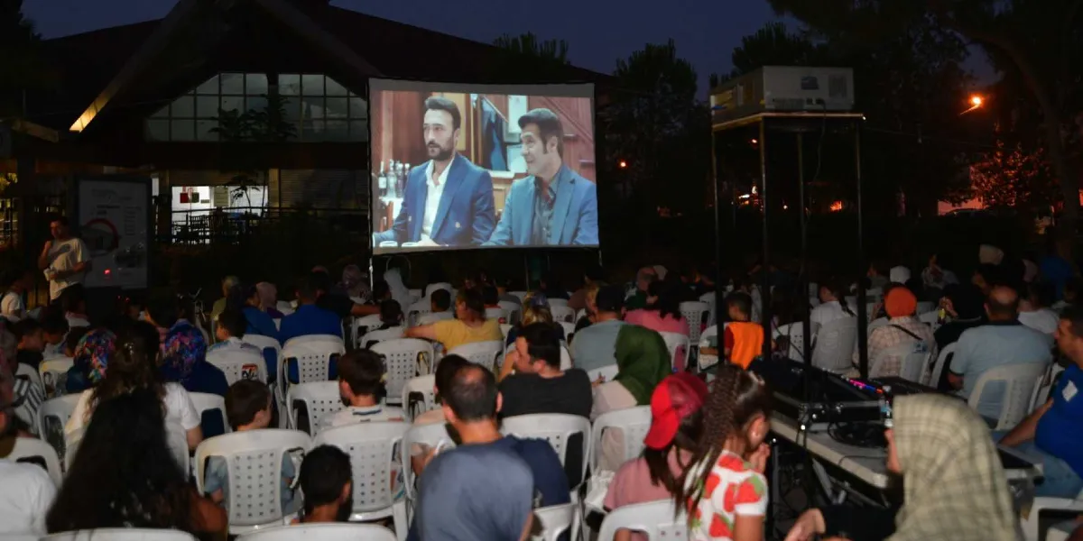 Açık havada, akşam saatlerinde kurulan büyük bir beyaz perde üzerinde film izleyen kalabalık bir izleyici grubu. İnsanlar, perdeye dönük şekilde dizilmiş beyaz plastik sandalyelerde oturuyor. Perdede, takım elbiseli iki erkek oyuncunun yer aldığı bir sahne yansıtılıyor. Sağ tarafta, yüksek bir platform üzerinde projeksiyon cihazı ve teknik ekipmanlar bulunuyor.