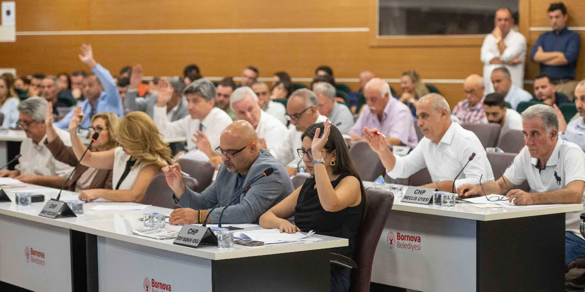 Bornova Belediyesi meclis salonunda düzenlenen toplantıda, katılımcıların büyük çoğunluğu ellerini kaldırarak oylamaya katılıyor. Masaların üzerinde Bornova Belediyesi logoları ve CHP Grup Başkan Vekili ile CHP Meclis Üyesi yazılı isimlikler yer alıyor. Ön planda, siyah kıyafetli bir kadın ve gri ceketli bir erkeğin de aralarında bulunduğu meclis üyeleri, önlerindeki evraklarla birlikte oturarak oylama sürecine dahil oluyor.
