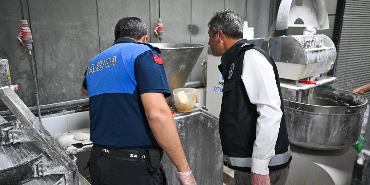 Zabıta üniformalı iki görevli, bir fırın imalathanesinde hamur yoğurma makinelerini ve üretim alanını denetliyor. Görevlilerden biri üzerinde 