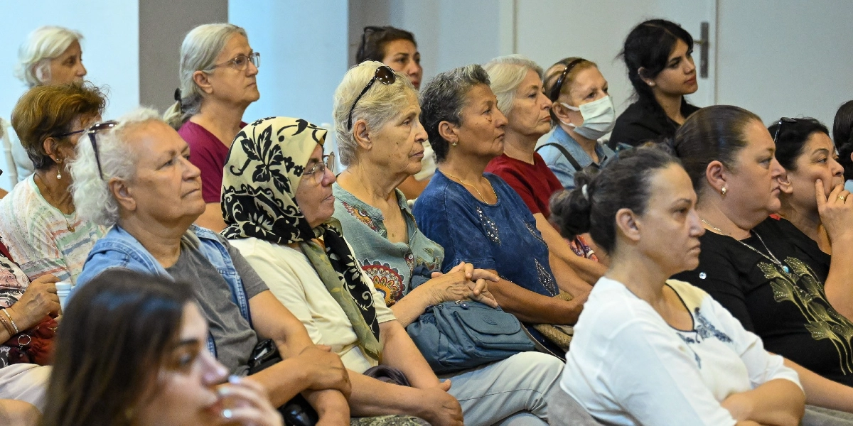 Bir konferans salonunda yan yana oturmuş, dikkatle dinleyen çoğunluğu orta yaş ve üzeri kadınlardan oluşan bir grup izleyici. Katılımcıların çoğu farklı renklerde kıyafetler giymiş, bazıları gözlük takmış, biri ise beyaz bir maske takıyor. İzleyiciler, önlerindeki bir konuşmacıyı veya sunumu takip eder bir ifadeyle karşıya bakıyor.