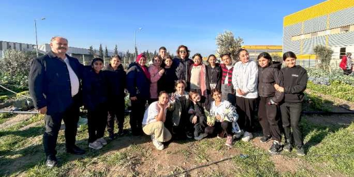 Açık havada, bir bahçede yan yana duran, aralarında yetişkinlerin de bulunduğu öğrenci grubu. Grubun sol tarafında lacivert ceketli bir erkek, diğerlerinde ise çoğunlukla günlük kıyafetler ve montlar var. Arka planda modern bir bina ve tarım alanı görülüyor.