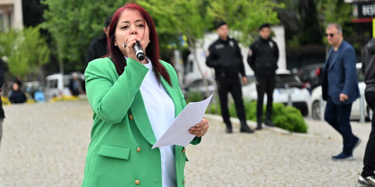 Kızıl saçlı bir kadın, açık havada elinde tuttuğu bir metni mikrofona okuyarak konuşma yapıyor. Üzerinde beyaz bir gömlek ve yeşil bir ceket bulunan kadının arkasında, güvenlik önlemi alan iki polis memuru ve yürüyen bir adam yer alıyor.
