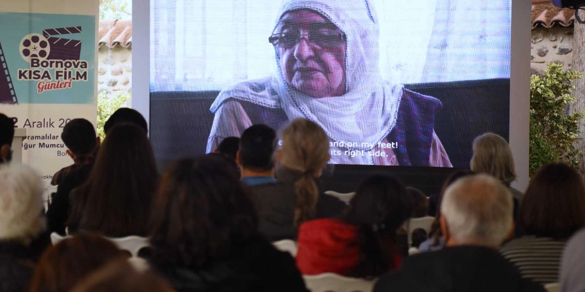 Açık havada düzenlenen bir film gösteriminde, izleyiciler arkadan görünürken dev ekranda gözlüklü ve başörtülü yaşlı bir kadının yakın çekim görüntüsü yer alıyor. Ekranın sol tarafında Bornova Kısa Film Günleri logosu ve 2 Aralık tarihini içeren bir bilgilendirme panosu bulunuyor.