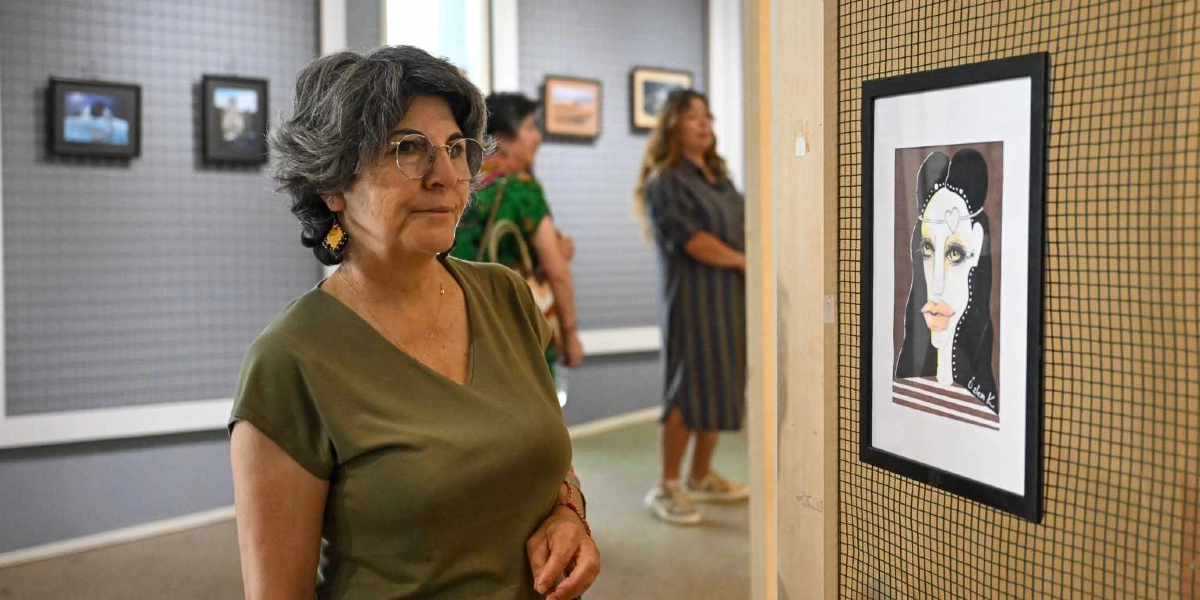Kısa gri saçlı, gözlüklü ve haki yeşili tişört giyen bir kadın, sanat galerisinde duvarda asılı olan, üzerinde stilize edilmiş bir kadın portresinin bulunduğu tabloyu dikkatle inceliyor. Arka planda galeriyi gezen diğer ziyaretçiler ve duvarda asılı başka tablolar yer alıyor.