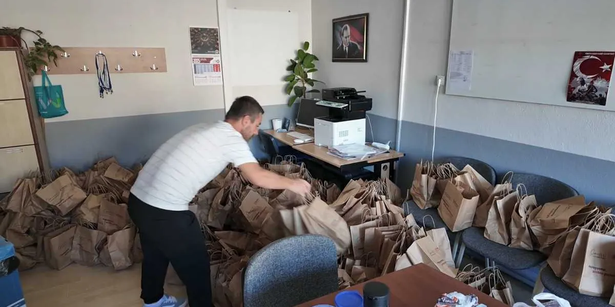 Ofis ortamında, üzerinde Bornova Belediyesi logosu bulunan çok sayıda kahverengi kağıt çantayı düzenleyen bir erkek görevli. Çantalar hem yerlerde hem de sandalyelerin üzerinde sıralanmış durumda. Arka planda bir çalışma masası, yazıcı, duvarda Atatürk portresi ve Türk bayrağı görseli yer alıyor.