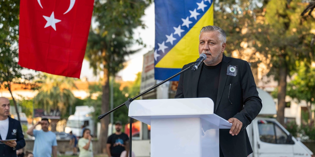 Açık havada, arkasında Türkiye ve Bosna-Hersek bayraklarının asılı olduğu bir kürsüde konuşma yapan, siyah ceketli ve siyah tişörtlü, gri saçlı bir adam. Adamın ceketinin yakasında Srebrenitsa soykırımını simgeleyen 
