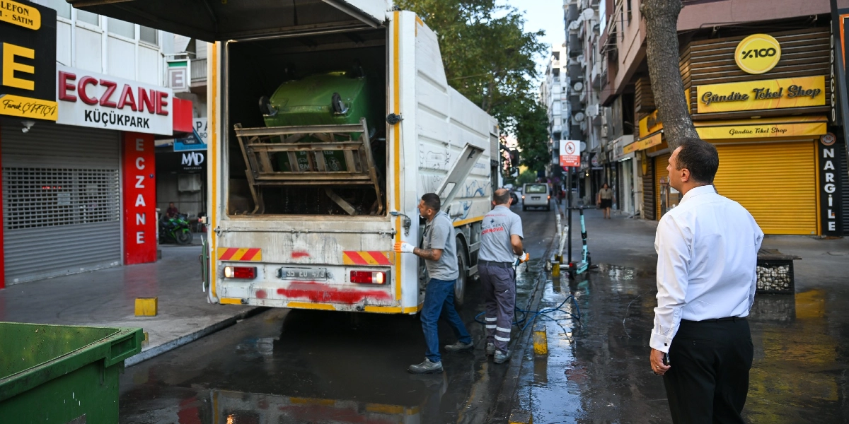 Bornova Belediye Başkanı Ömer Eşki, sabah saatlerinde bir şehir caddesinde temizlik çalışmalarını denetliyor. Belediye personeli, çöp kamyonunun mekanizmasını kullanarak yeşil bir çöp konteynerini boşaltırken, Başkan Eşki çalışmaları dikkatle izliyor. Cadde üzerinde eczane ve tekel bayii gibi dükkanların bulunduğu, ıslak zeminli bir sokak ortamı görülüyor.