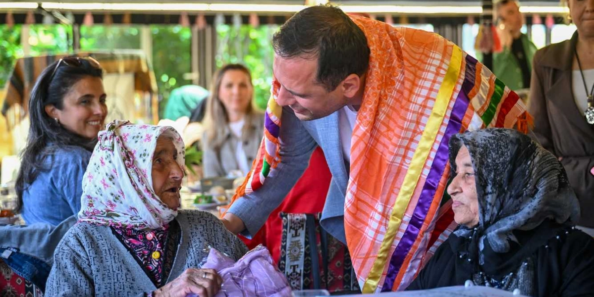 Omuzlarında renkli, geleneksel bir şal bulunan bir erkek, Anneler Günü etkinliğinde iki yaşlı kadının yanına eğilerek onlarla sohbet ediyor. Kadınlardan biri çiçekli bir başörtüsü takarken, diğeri koyu renkli bir eşarp kullanıyor. Arka planda ise etkinliğe katılan diğer insanlar gülümseyerek onları izliyor.