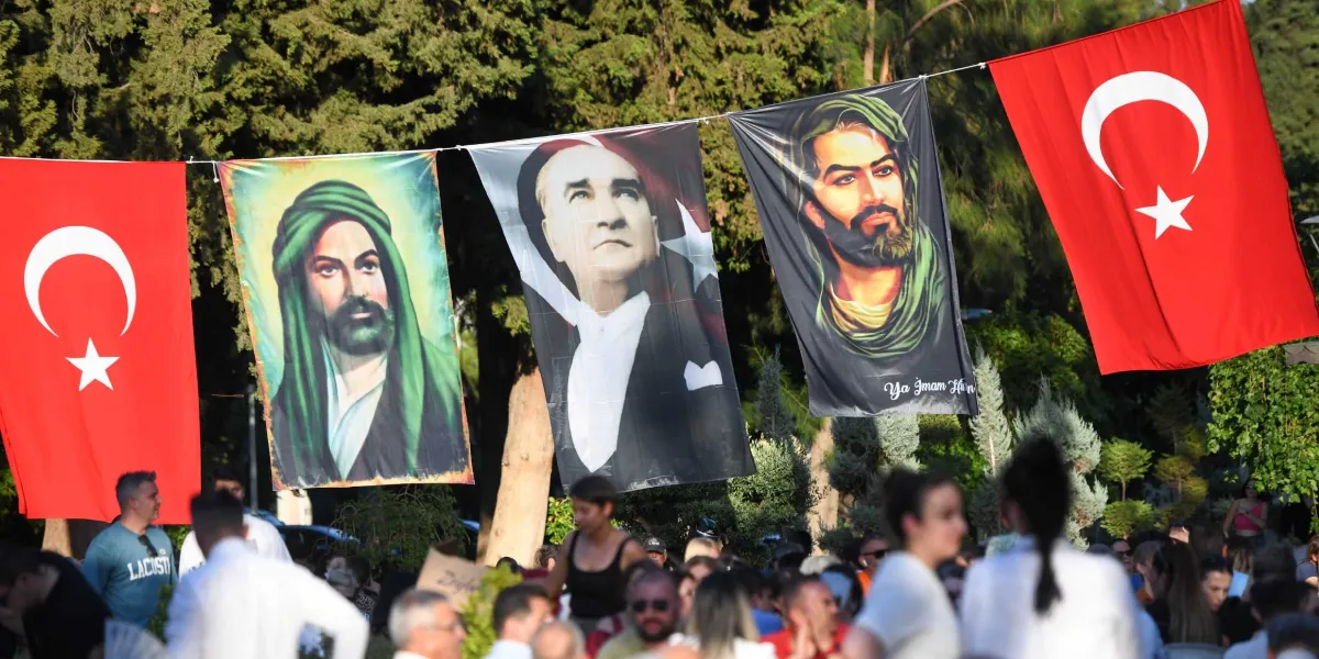 Açık havada, ağaçların önünde bir ipe yan yana asılmış dört büyük afiş ve kalabalık bir insan grubu. En solda ve en sağda Türk bayrakları yer alıyor. Ortadaki iki afişten birinde Mustafa Kemal Atatürk, diğerlerinde ise yeşil sarıklı ve sakallı dini figürlerin portreleri bulunuyor. İnsanlar bu afişlerin altında bir etkinlik için toplanmış durumda.